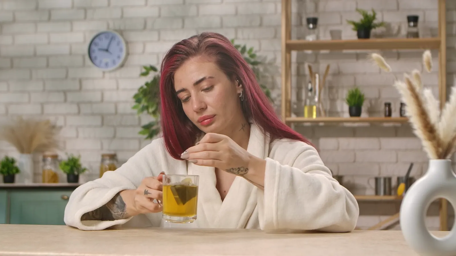 A woman in a robe at her kitchen table, looking down thoughtfully at a cup of tea